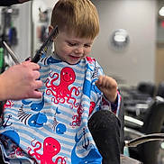 Eli is registered to the contest to win money with this photo: barbershop, boy, cape, chair, child, clothing, comb, cute, haircut, haircutting, hairdresser, hairstyle, happy, indoor, kid, person, scissors, short_hair, sitting, smile