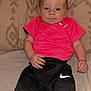 Ralphie is registered to the contest to win money with this photo: baby, child, infant, couch, clothing, pink_shirt, black_pants, nike_logo, face, hands, indoor, portrait, seated, young_child, fabric, patterned_cushion, expression, skin, person, flash_photo