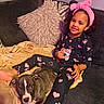 child, dog, puppy, couch, blanket, pillow, smile, pajamas, heart_pattern, headband, curly_hair, indoor, cozy, relaxing, friendship, pet, happy, living_room, comfort, cute
