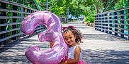 Eliana joined the competition — help win amazing prizes! balloon, bridge, celebration, child, curly_hair, footwear, girl, greenery, happy, nature, number_three, outdoor, park, pink_dress, portrait, ribbon, smiling, summer, sunlight, wooden_floor