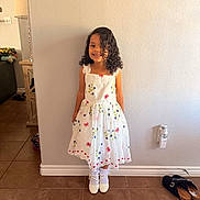 Eliana joined the competition — help win amazing prizes! child, girl, curly_hair, white_dress, floral_pattern, smiling, standing, indoor, tile_floor, socks, shoes, wall, light_switch, picture_frame, home_interior, footwear, lamp, furniture, happy, portrait