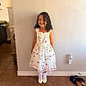 child, girl, curly_hair, white_dress, floral_pattern, smiling, standing, indoor, tile_floor, socks, shoes, wall, light_switch, picture_frame, home_interior, footwear, lamp, furniture, happy, portrait