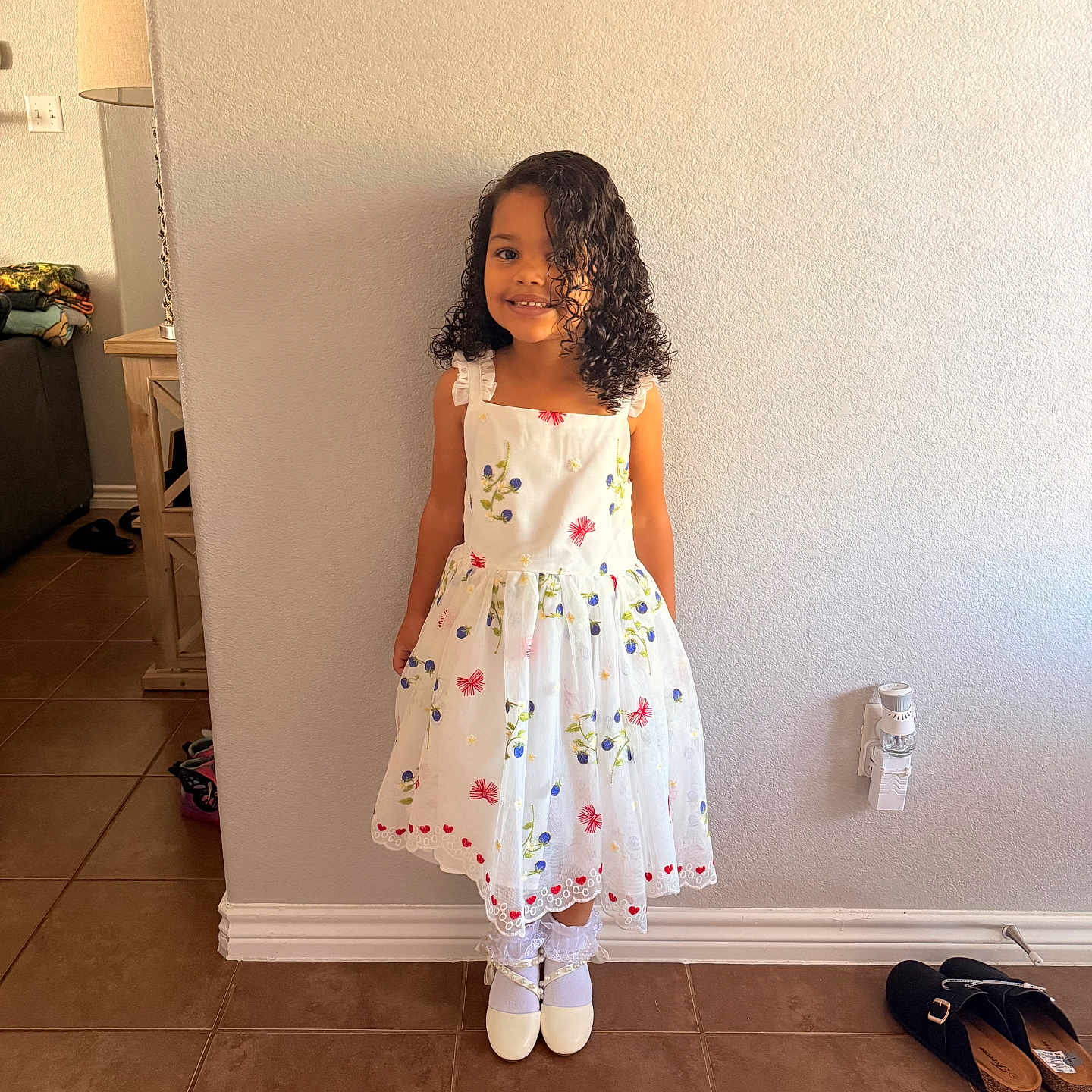 Eliana joined the competition — help win amazing prizes! child, girl, curly_hair, white_dress, floral_pattern, smiling, standing, indoor, tile_floor, socks, shoes, wall, light_switch, picture_frame, home_interior, footwear, lamp, furniture, happy, portrait