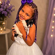 Eliana joined the competition — help win amazing prizes! child, girl, puppy, dog, braided_hair, purple_bow, white_dress, smiling, hugging, indoor, portrait, cute, happy, pet, flowers, table, decor, lights, beads, toddler