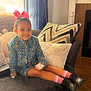 Eliana joined the competition — help win amazing prizes! child, girl, smiling, pink_bow, hair_bun, denim_jacket, patterned_jacket, pink_socks, black_shoes, couch, throw_pillow, living_room, curtain, balloons, keyboard, plush_toy, fireplace, sitting, legs, toy