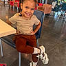 child, girl, smiling, portrait, sitting, chair, cafe, indoor, braids, hair_bows, brown_hair, shirt, skirt, leggings, socks, sneakers, crossed_legs, waiting_area, wooden_table, happy