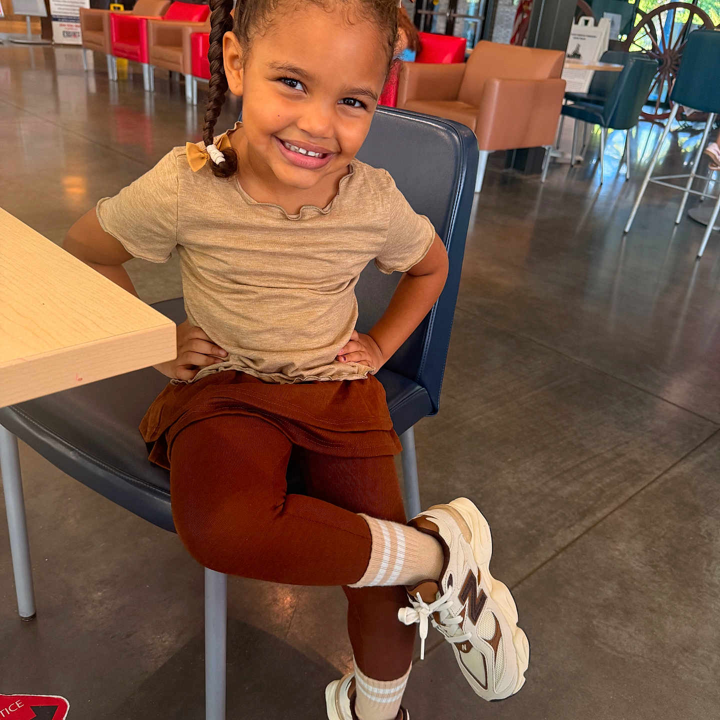 Eliana is registered to the contest to win money with this photo: child, girl, smiling, portrait, sitting, chair, cafe, indoor, braids, hair_bows, brown_hair, shirt, skirt, leggings, socks, sneakers, crossed_legs, waiting_area, wooden_table, happy