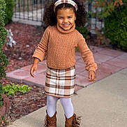 Eliana joined the competition — help win amazing prizes! child, girl, smile, curly_hair, headband, brown_sweater, plaid_skirt, white_tights, brown_boots, fringe, outdoor, sidewalk, garden, fence, happy, fashion, portrait, standing, person, daylight