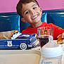 baby_bottle, blue_booth, boy, bread, child, dining, food, fork, glass, happy, indoor, kid, pink_wall, playful, police_car, red_shirt, smile, soda, table, toy