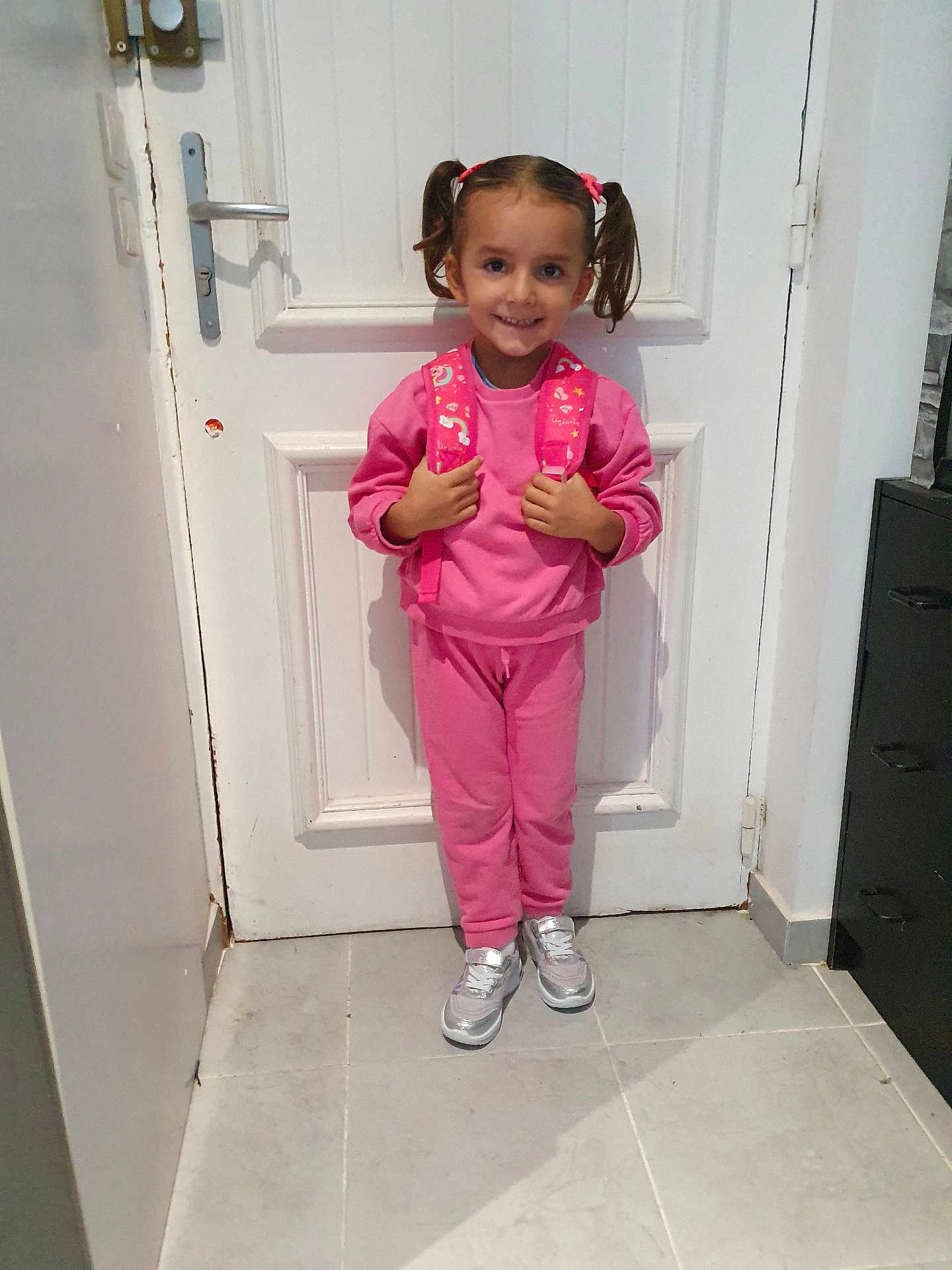 Valentina a rejoint le concours — aidez-le/la à gagner de superbes lots ! child, girl, pink_clothing, backpack, pigtails, smile, door, white_door, indoor, tile_floor, sneakers, silver_shoes, standing, happy, person, hair_ties, casual_wear, young_child, portrait, home_interior