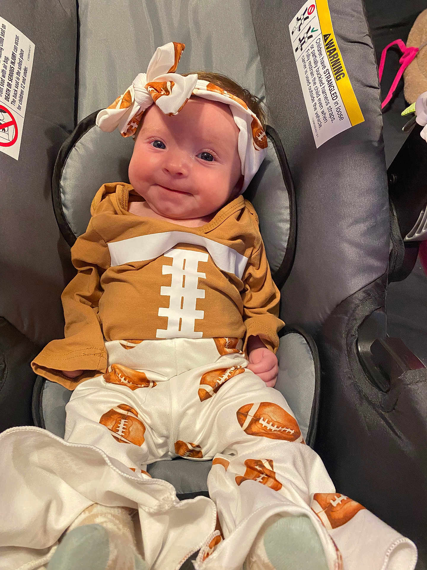 Luna is registered to the contest to win money with this photo: baby, infant, car_seat, football, headband, smile, clothing, cute, child, seat, sitting, face, portrait, indoor, young, happy, apparel, patterned_pants, soft_lighting, comfortable