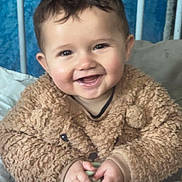 Zackary a rejoint le concours — aidez-le/la à gagner de superbes lots ! baby, bars, blue_background, brown_clothing, child, cozy, crib, cute, face, fuzzy_sweater, hands, happy, indoor, infant, person, portrait, sitting, smiling, toy, young_child