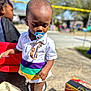 blurred_background, casual_wear, child, colorful_clothing, curious, daytime, outdoor, pacifier, person, playing, portrait, shoes, shorts, snack_box, standing, summer, sunlight, toddler, toy_wagon, young_child