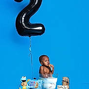 Chad is registered to the contest to win money with this photo: balloon, birthday, blue_background, celebration, child, cookie, cookie_jar, cookie_monster, curious, food, glass_bowl, milk, number_2, packaging, reflection, sitting, snack, table, toddler, toy