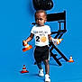 Chad is registered to the contest to win money with this photo: balloon, basketball_graphic, black_shorts, blue_background, boy, child, confident, director_chair, indoor, number_2, playful, portrait, sports_theme, studio, toddler, traffic_cones, walking, white_shoes, white_tshirt, young_child