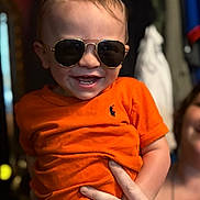 Brayden is registered to the contest to win money with this photo: toddler, child, sunglasses, orange_shirt, smile, happy, person, hand, indoor, clothing, face, portrait, cute, baby, fashion, accessory, holding, ring, tattoo, fun