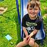 child, boy, smiling, slide, playground, outdoor, grass, sunlight, casual_clothing, shorts, shoes, black_tshirt, toy, happy, summer, play, young, fun, nature, recreation