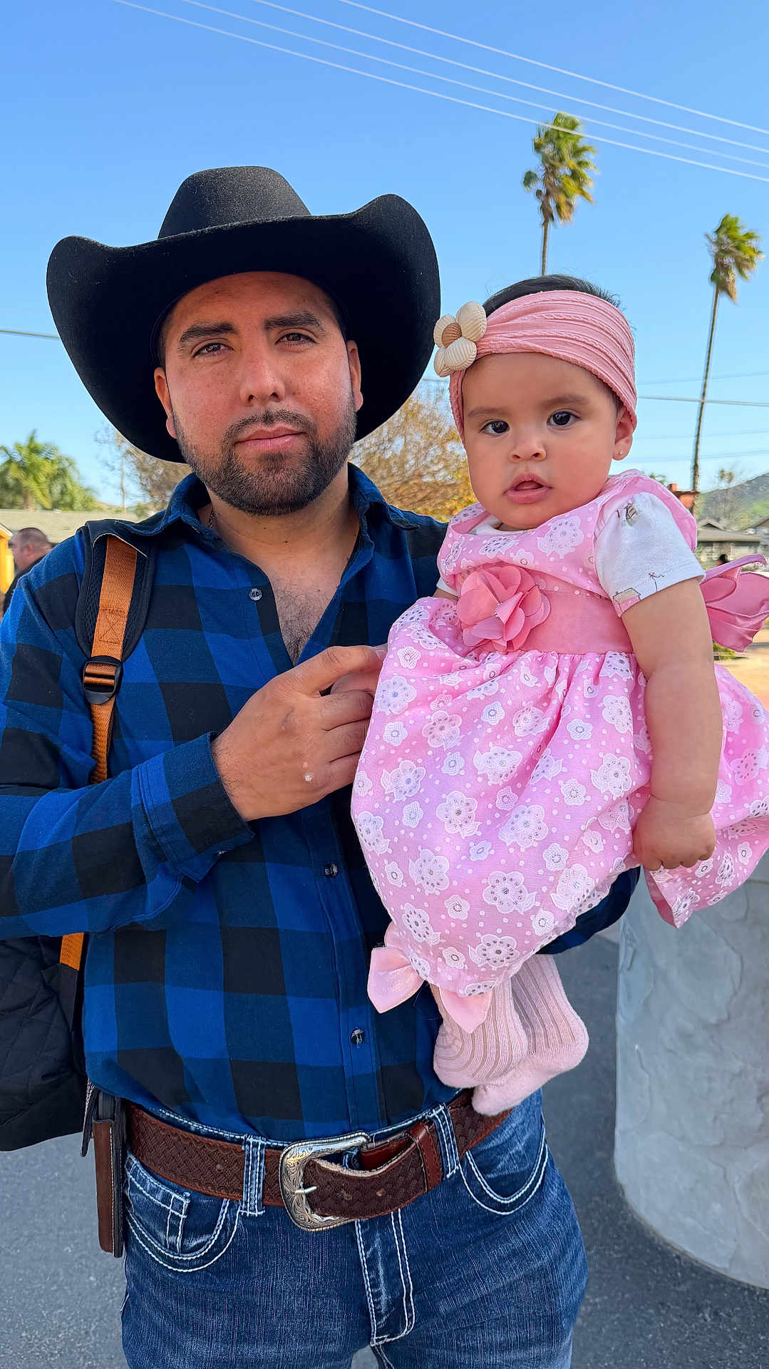 Lesly is registered to the contest to win money with this photo: man, baby, child, cowboy_hat, pink_dress, headband, outdoor, daylight, portrait, smile, beard, jeans, belt, backpack, person, sky, trees, palm_trees, holding, family