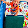 baby, child, smiling, happy, playpen, indoor, toy, colorful_balls, alphabet_mat, cushion, orange_shirt, shorts, standing, portrait, face, eyes, teeth, playroom, infant, photography