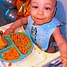 baby, child, high_chair, eating, food, pasta, mashed_food, bib, blue_bib, smiling, face, portrait, indoor, birthday, balloons, party, colorful, table, messy_mealtime, seated