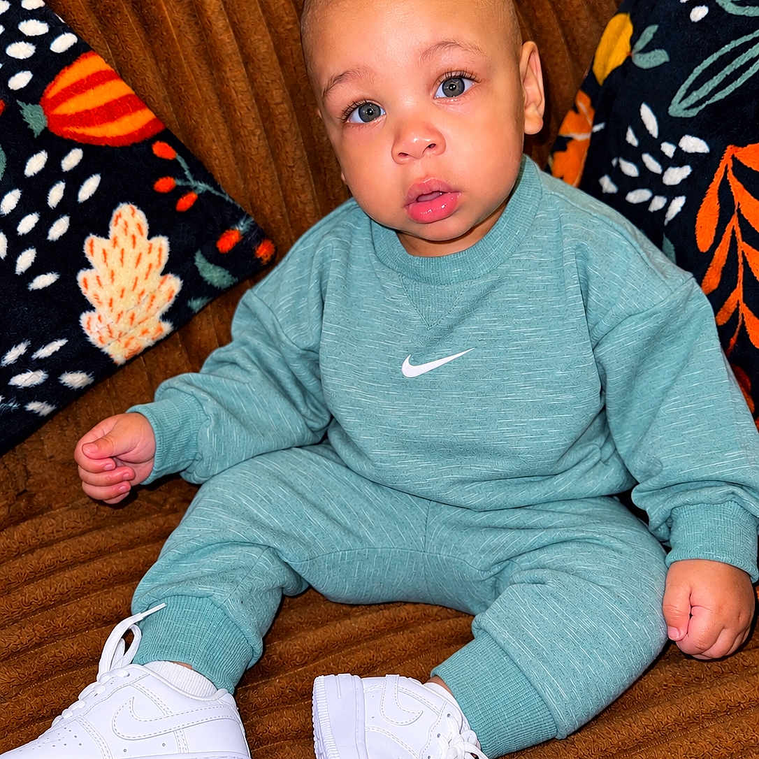 Braylen is registered to the contest to win money with this photo: baby, child, infant, sitting, portrait, blue_eyes, wide_eyed, mouth_open, tracksuit, sweatshirt, sneakers, nike, chair, couch, blanket, patterned_fabric, indoor, fashion, closeup, smiling