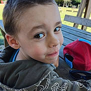 Tristan a rejoint le concours — aidez-le/la à gagner de superbes lots ! child, boy, face, smile, outdoor, park, picnic_table, hoodie, cap, playground, tree, daylight, casual_clothing, young, person, bench, nature, happy, head, portrait