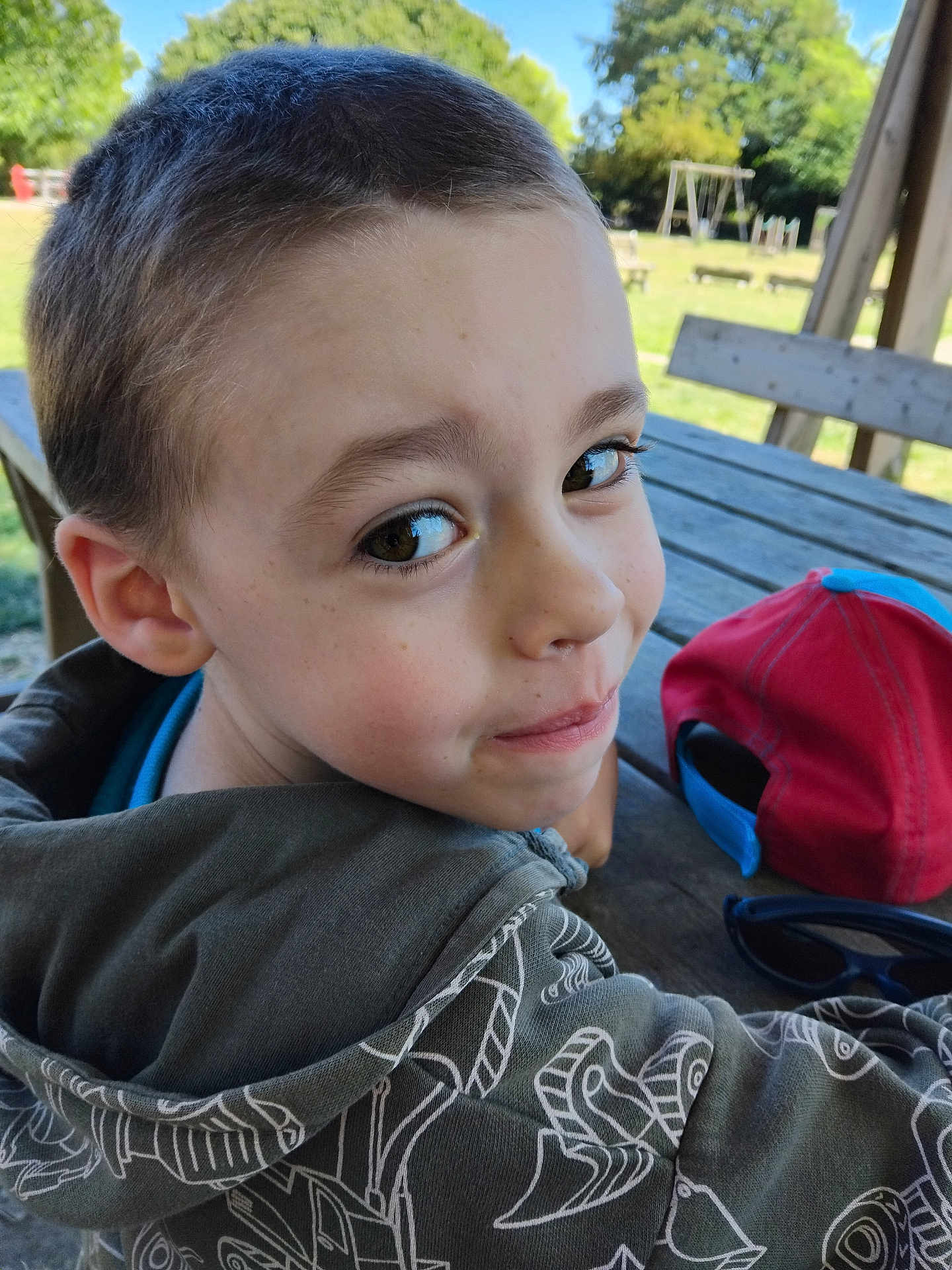 Tristan a rejoint le concours — aidez-le/la à gagner de superbes lots ! child, boy, face, smile, outdoor, park, picnic_table, hoodie, cap, playground, tree, daylight, casual_clothing, young, person, bench, nature, happy, head, portrait