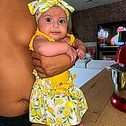 Alma a rejoint le concours — aidez-le/la à gagner de superbes lots ! baby, child, person, shirtless, headband, lemon_pattern, yellow_clothing, arm, hand, kitchen, mixer, wooden_counter, television, living_room, brick_wall, couch, foot, table, indoor, holding