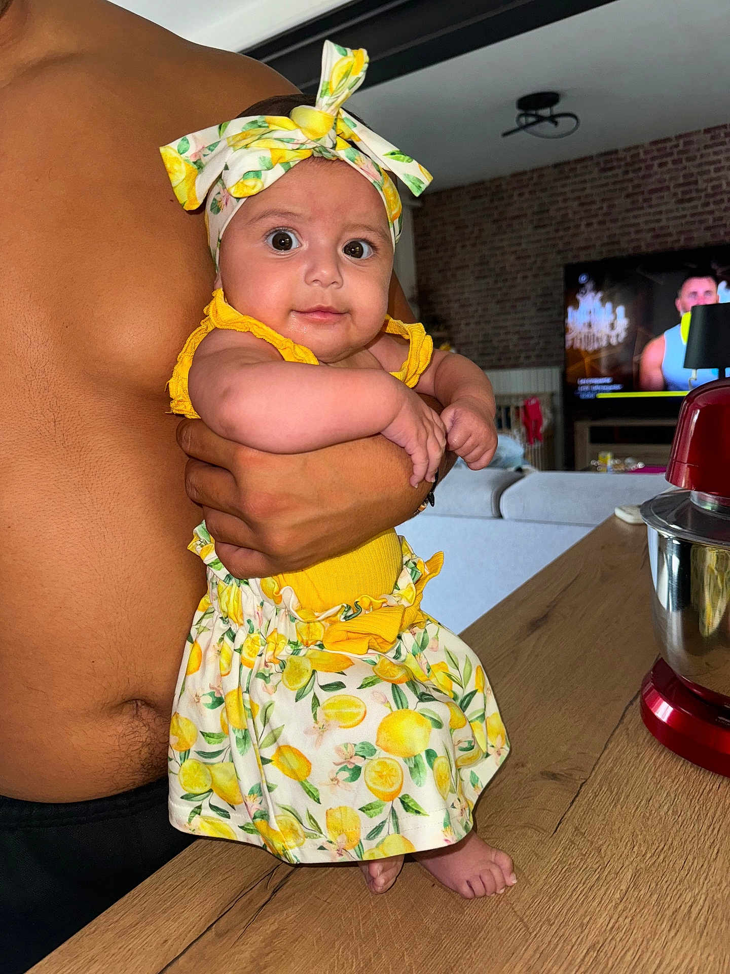 Alma a rejoint le concours — aidez-le/la à gagner de superbes lots ! baby, child, person, shirtless, headband, lemon_pattern, yellow_clothing, arm, hand, kitchen, mixer, wooden_counter, television, living_room, brick_wall, couch, foot, table, indoor, holding