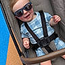 baby, child, infant, stroller, sunglasses, smiling, happy, portrait, outdoor, seated, harness, seatbelt, blue_clothing, patterned_clothing, fair_skin, brown_hair, playground, carpet, closeup, adorable