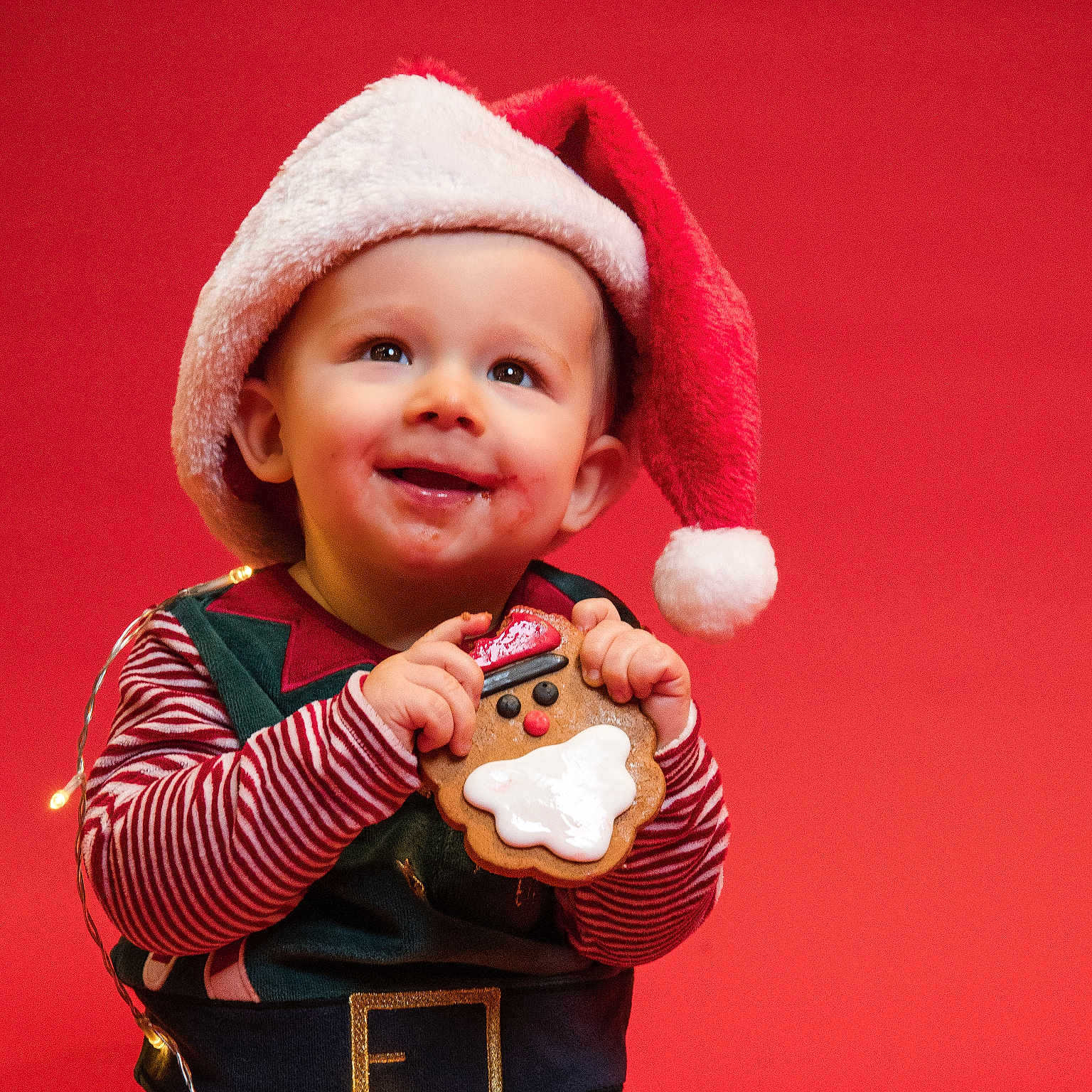 Callahan is registered to the contest to win money with this photo: celebration, child, christmas, cute, festive, festive_clothing, gingerbread_cookie, happy, holiday, holiday_season, indoors, joyful, portrait, red_background, santa_hat, smiling, string_lights, striped_shirt, toddler, winter