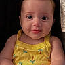baby, infant, yellow_clothing, romper, cute, smiling, face, child, portrait, hands, sitting, indoor, seat, car_seat, curly_hair, skin, expression, young_child, person, closeup