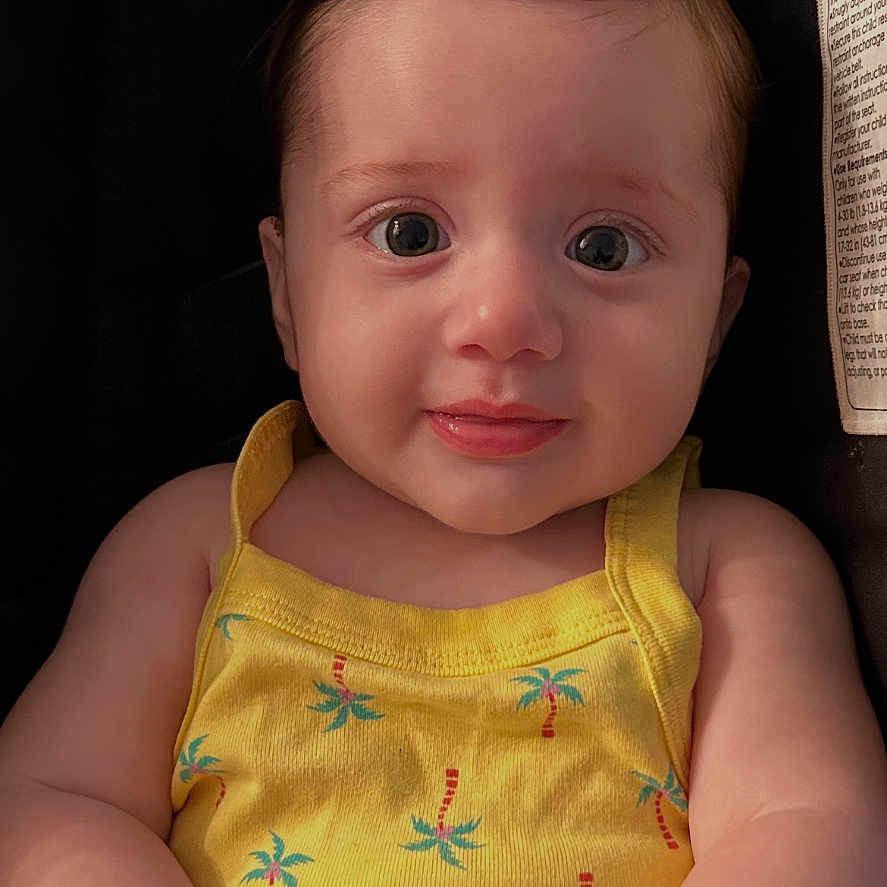 Anna is registered to the contest to win money with this photo: baby, infant, yellow_clothing, romper, cute, smiling, face, child, portrait, hands, sitting, indoor, seat, car_seat, curly_hair, skin, expression, young_child, person, closeup