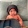 Elijah a rejoint le concours — aidez-le/la à gagner de superbes lots ! baby, child, guitar, acoustic_guitar, hat, knit_cap, cushion, green_cushion, indoor, musical_instrument, person, curious, seated, looking_away, skin, arms, strings, wood, furniture, casual