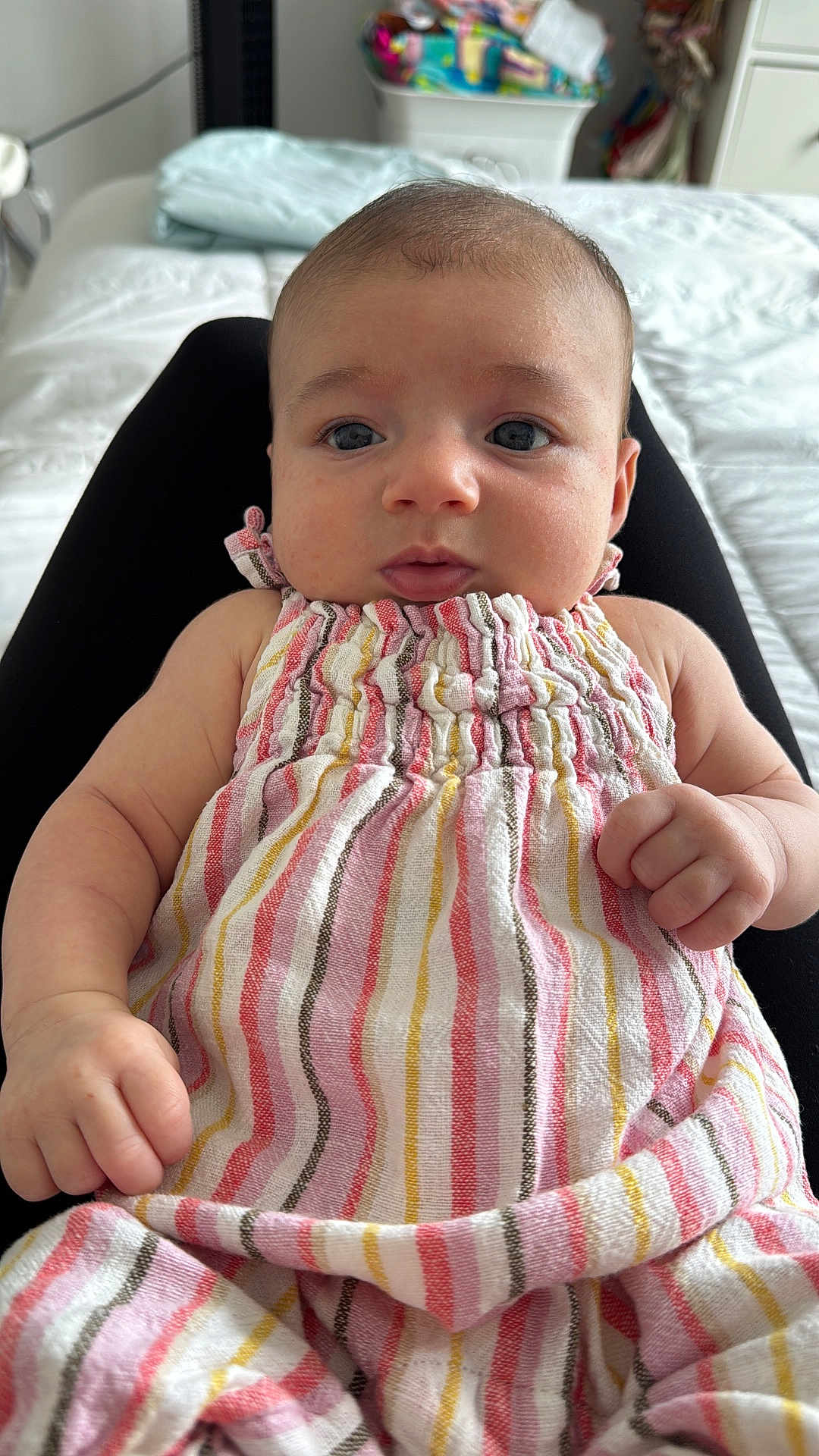 Bellamy is registered to the contest to win money with this photo: baby, infant, child, dress, striped_clothing, person, face, eyes, hand, fist, lap, bed, blanket, indoor, furniture, soft, curious, closeup, skin, cute
