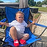 blue_chair, casual_clothing, child, curly_hair, daytime, deck, expression, fence, grass, gravel_path, outdoor, person, red_socks, shorts, sitting, sunny, trees, vehicle, white_tshirt, wooden_deck