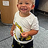 background_objects, carpet, child, clothing, curly_hair, drum, excavator_print, expression, floor, footwear, happy, indoor, person, playful, shorts, smiling, standing, toddler, white_tshirt, yellow_shoes