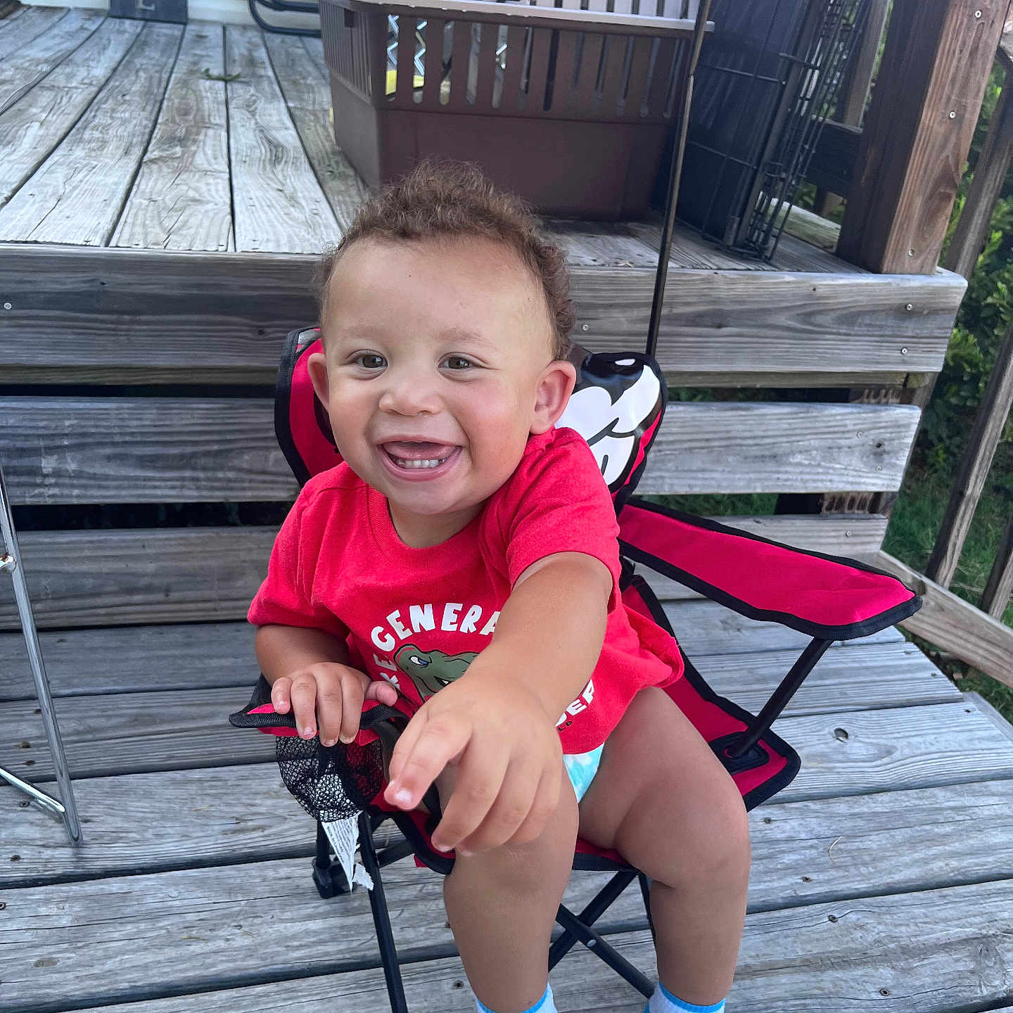 Kairi joined the competition — help win amazing prizes! toddler, child, smiling, red_shirt, chair, folding_chair, wooden_deck, outdoor, umbrella, happy, baby, socks, person, seat, wood, hand, face, grass, balcony, railing