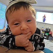 Persephone is registered to the contest to win money with this photo: baby, infant, child, close_up, hand, fingers, headband, bow, clothing, living_room, television, window, furniture, person, bed, blanket, home, indoor, face, cute