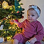 baby, child, infant, christmas_tree, ornament, holiday, decorations, pink_clothing, headband, indoors, smiling, cute, festive, holiday_lights, toy, sitting, face, person, celebration, seasonal
