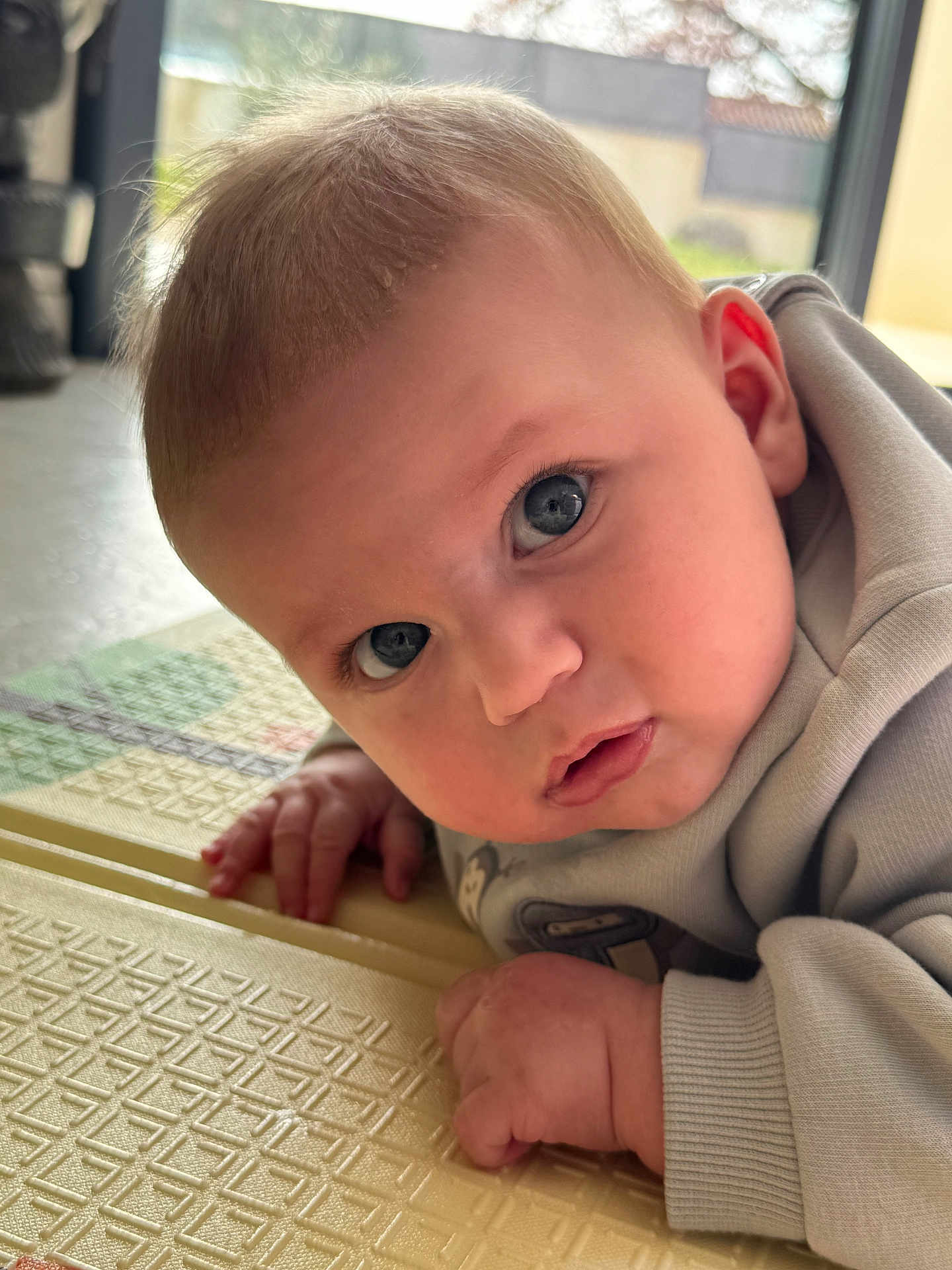 Louka participe au concours pour gagner de l'argent avec cette photo : baby, infant, blue_eyes, close_up, face, hand, playmat, textured_mat, indoors, window, natural_light, sweater, cheek, gaze, portrait, adorable, skin, ear, lying_down, curious_expression