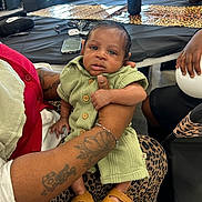 Koeven is registered to the contest to win money with this photo: baby, child, person, tattoo, hand, green_clothing, leopard_print, table, sunglasses, smartphone, sandals, indoor, seated, holding, furniture, decor, adult, skin, face, curious