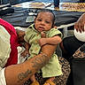 baby, child, person, tattoo, hand, green_clothing, leopard_print, table, sunglasses, smartphone, sandals, indoor, seated, holding, furniture, decor, adult, skin, face, curious