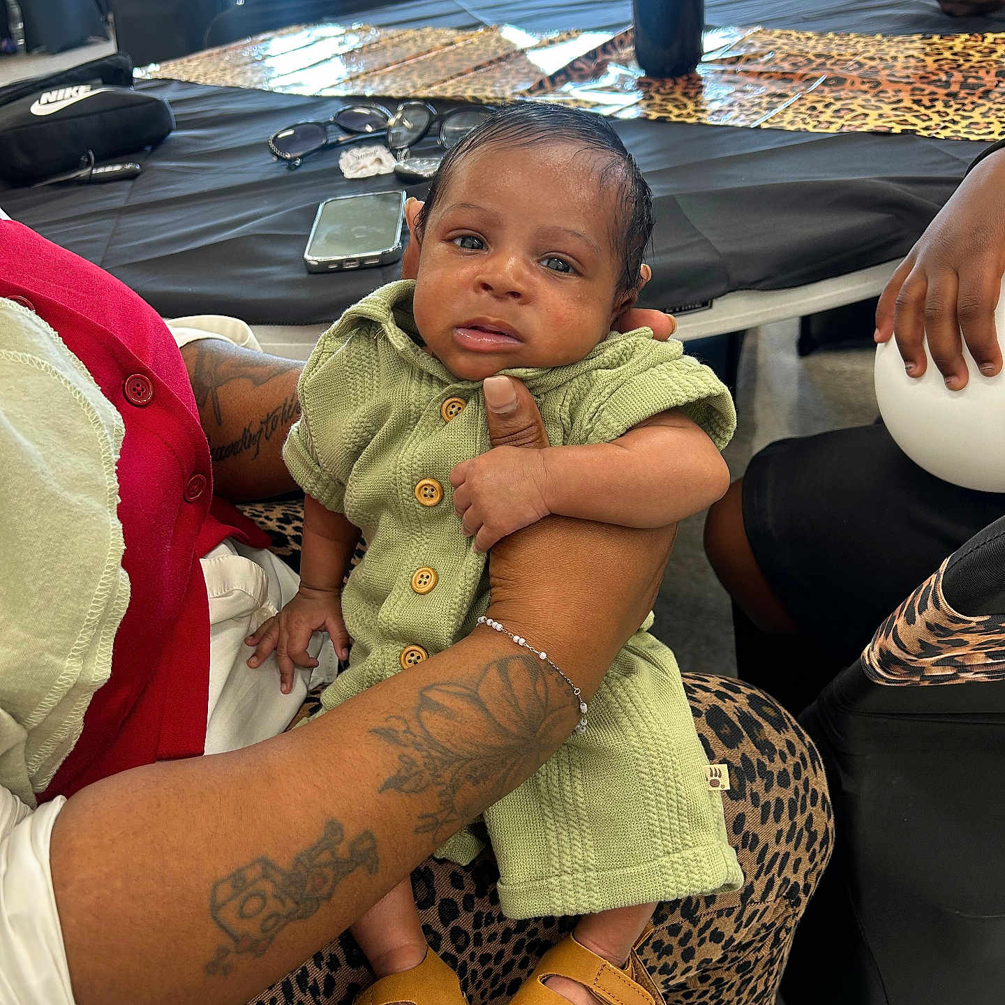 Koeven is registered to the contest to win money with this photo: baby, child, person, tattoo, hand, green_clothing, leopard_print, table, sunglasses, smartphone, sandals, indoor, seated, holding, furniture, decor, adult, skin, face, curious