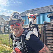 Sky joined the competition — help win amazing prizes! man, dog, sunglasses, hat, outdoor, smiling, tattoo, backpack, goggles, pet, sunlight, casual_clothing, grass, fence, building, blue_sky, happy, earrings, beard, portrait