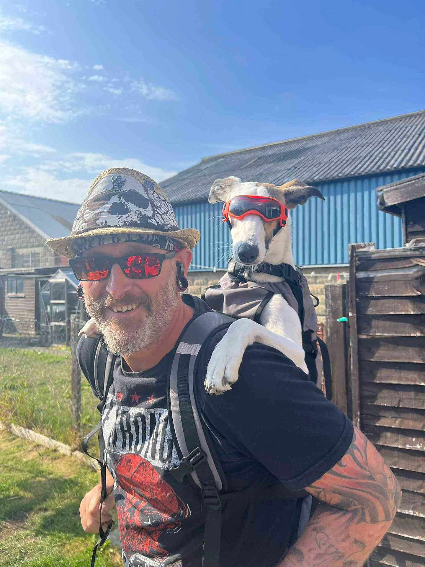Sky joined the competition — help win amazing prizes! man, dog, sunglasses, hat, outdoor, smiling, tattoo, backpack, goggles, pet, sunlight, casual_clothing, grass, fence, building, blue_sky, happy, earrings, beard, portrait