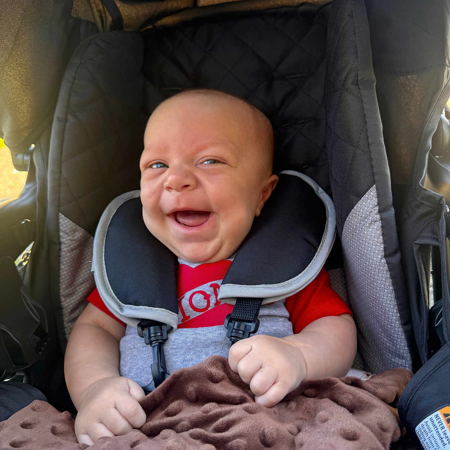 Josiah joined the competition — help win amazing prizes! baby, smiling, car_seat, blanket, infant, child, seatbelt, happy, portrait, cute, person, red_clothing, black_seat, soft_texture, indoors, safety, young_child, head_support, baby_clothes, infant_happiness
