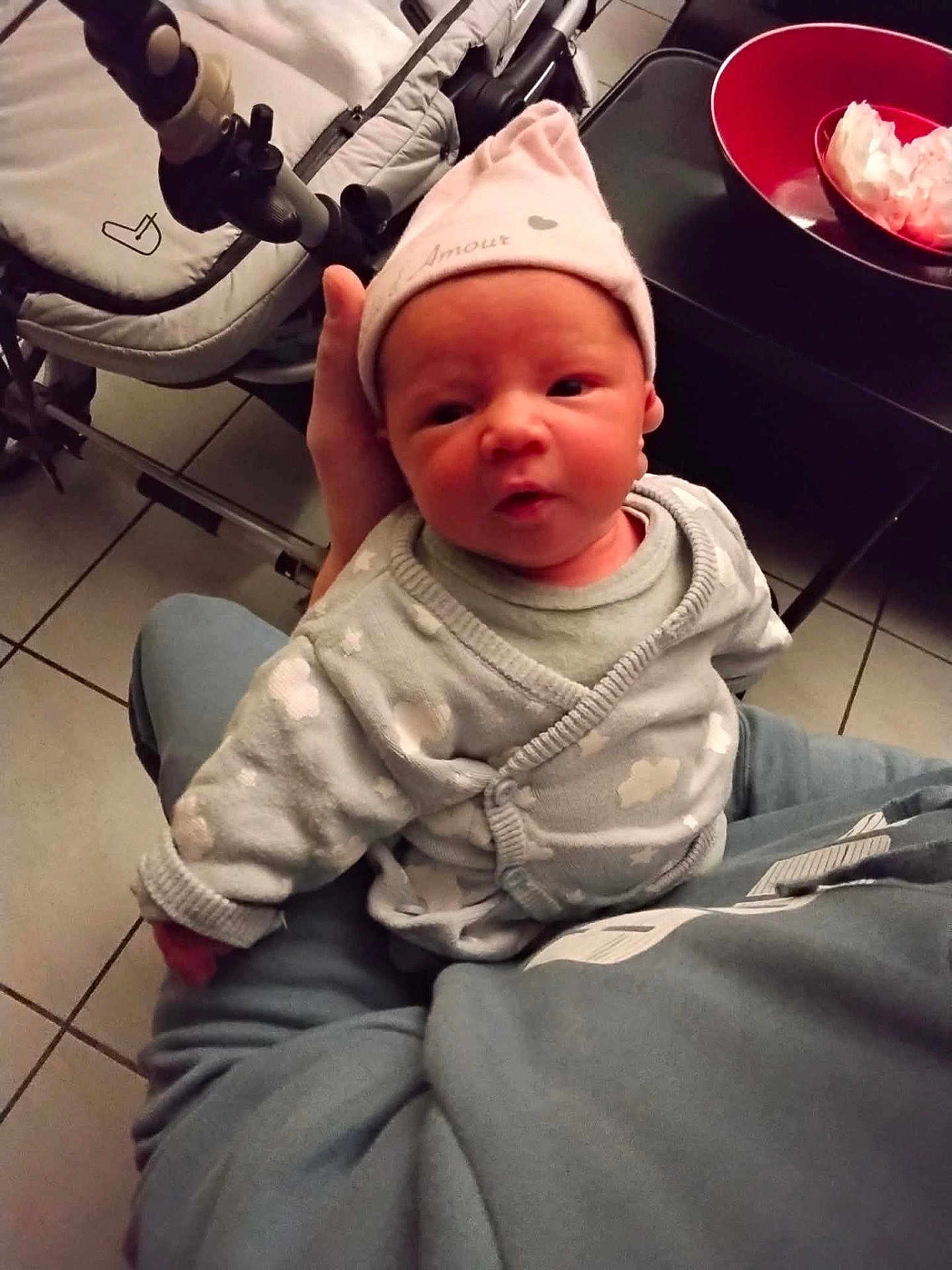 Joy a rejoint le concours — aidez-le/la à gagner de superbes lots ! baby, newborn, hat, onesie, stroller, lap, hand, tile_floor, red_bowl, bowl, indoor, person_holding, cute, portrait, seated, cozy, soft_clothing, expression, household, parent_lap