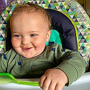 Luke joined the competition — help win amazing prizes! baby, child, high_chair, spoon, green_shirt, dinosaur_print, smile, face, hand, food, kitchen, indoor, person, toddler, cute, happy, messy, feeding, seat, portrait