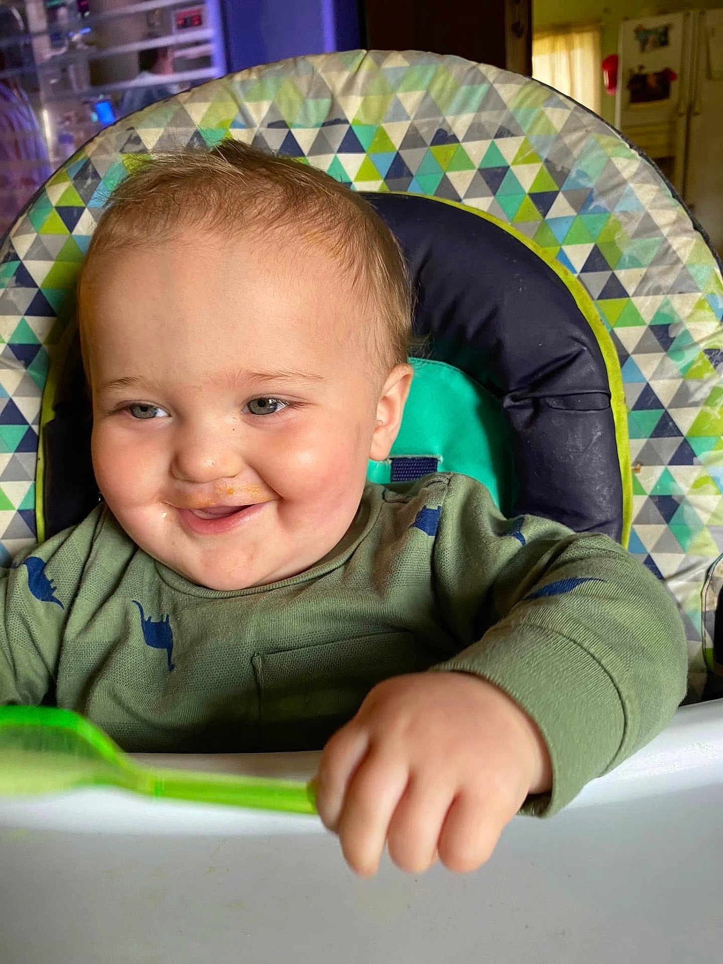 Luke joined the competition — help win amazing prizes! baby, child, high_chair, spoon, green_shirt, dinosaur_print, smile, face, hand, food, kitchen, indoor, person, toddler, cute, happy, messy, feeding, seat, portrait